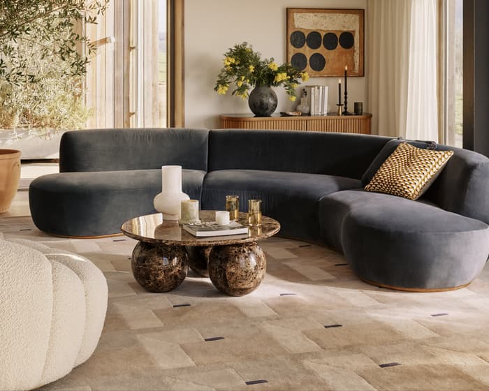 A curved dark-gray sofa sits on a patterned rug, centered by a round marble coffee table with decorative objects, in a modern living room featuring large windows, a wooden ceiling, and a spherical chandelier.