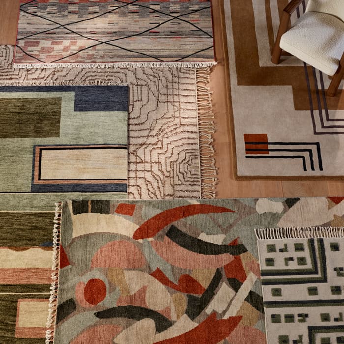 Several patterned rugs displaying abstract geometric designs lie on a wooden floor, with a cushioned wooden chair partially visible in the top-right corner.