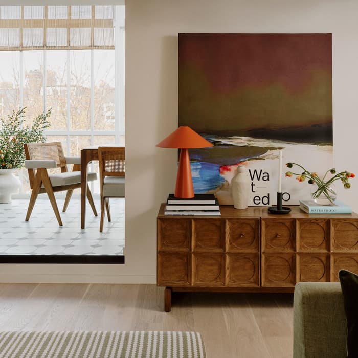 Wooden cabinet with orange lamp, books, vase, and abstract art; situated in a modern living room with patterned flooring and green couch. Visible dining area with wooden chairs and large windows. Text: "Wated wp."