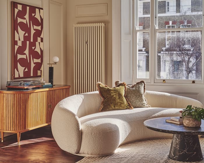 A curvy white sofa adorned with pillows sits in a sunlit living room near a black marble coffee table, sideboard with lamp, record player, and abstract artwork; large windows and radiator behind.