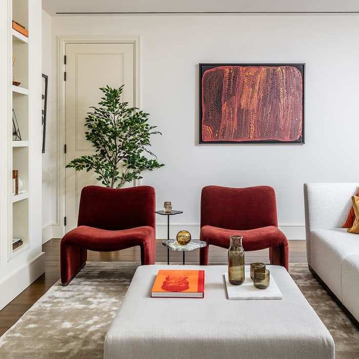 Two red chairs sit next to a small table and a plant in a modern living room; an abstract painting hangs on the white wall above, with books and glassware on a coffee table.