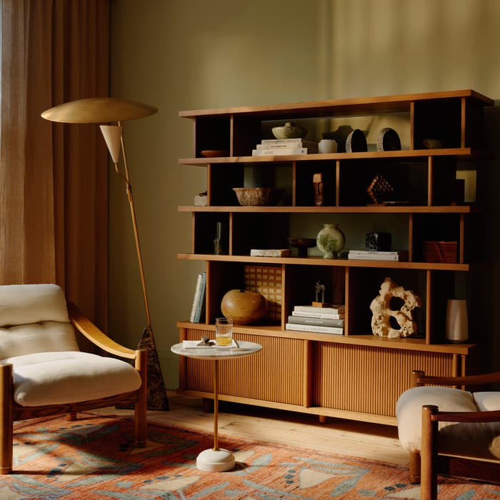 Wooden armchair with white cushion sits beside a tall, golden floor lamp and marble side table holding a glass; against a large, wooden bookshelf with various decorative objects in a cozy living room.