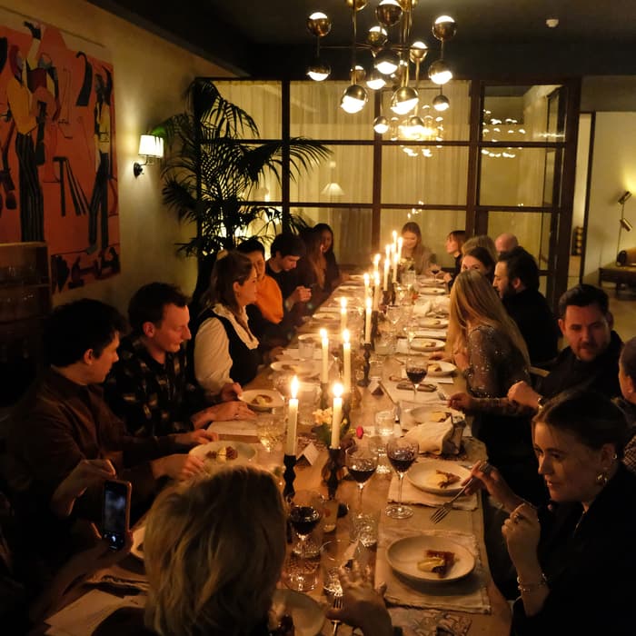 People sitting around a long dining table, enjoying a meal by candlelight in a warmly-lit, modern dining room with large windows, a contemporary chandelier, and a colorful wall painting.