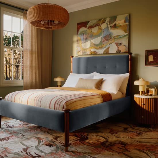 Bed adorned with striped bedding in a warmly lit bedroom. On the nightstand is a lamp, abstract painting above the bed, rug on the wooden floor, and large windows with beige curtains.