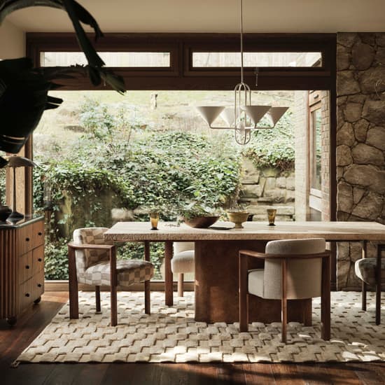 A dining table surrounded by cushioned chairs with a pendant light overhead in a stone-walled room, featuring large windows, green foliage outside, a patterned rug, and wooden furniture.