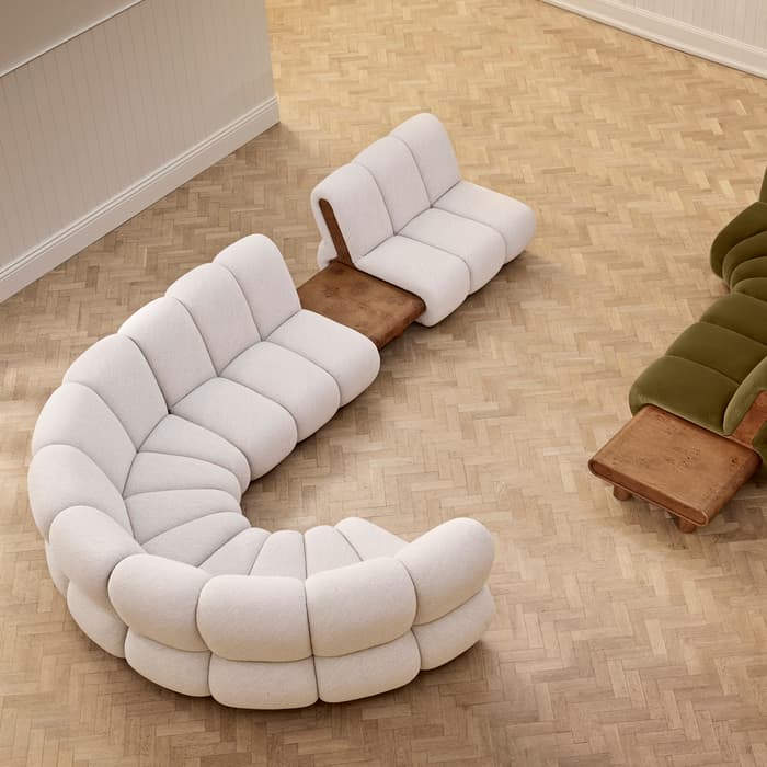 Curved, plush, sectional sofas—one white, one green—are arranged in a spacious, parquet-floored room, connected by wooden side tables. The room features white-paneled wainscoting.