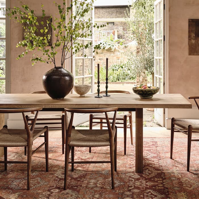 A wooden dining table with chairs holds a vase with branches, candlesticks, and a bowl. It's situated in an elegant room with large windows and two chandeliers, overlooking a garden.