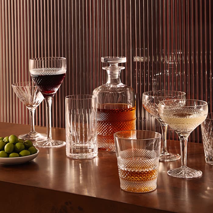 Crystal glasses filled with red wine, whiskey, champagne, and a carafe on a wooden counter with a bowl of green olives, set against a background of vertical blinds.