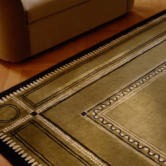 A beige sofa stands on a parquet wooden floor beside a green and black patterned rug featuring geometric designs and a rectangular border.