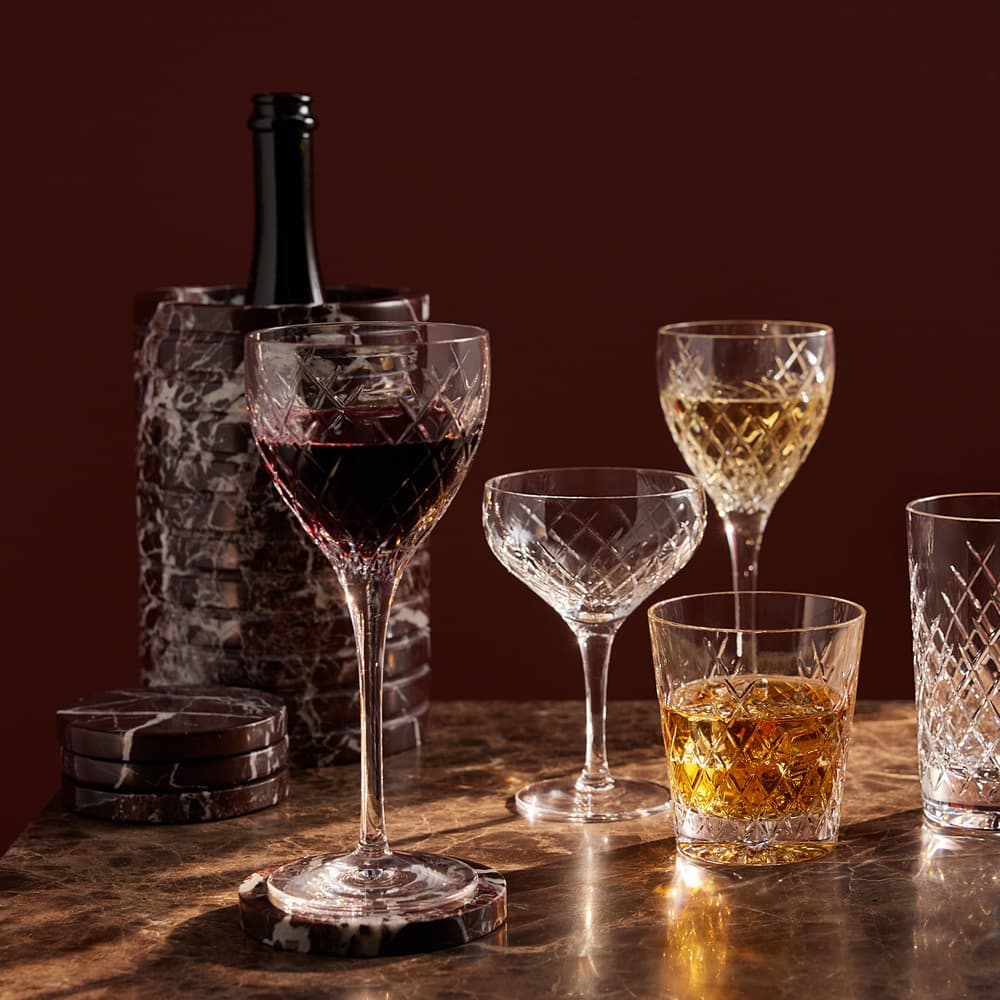 Crystal glasses filled with various drinks are arranged on a marble surface, with a dark wine bottle partially visible inside a matching ice bucket; coasters lie nearby in a softly lit, elegant setting.