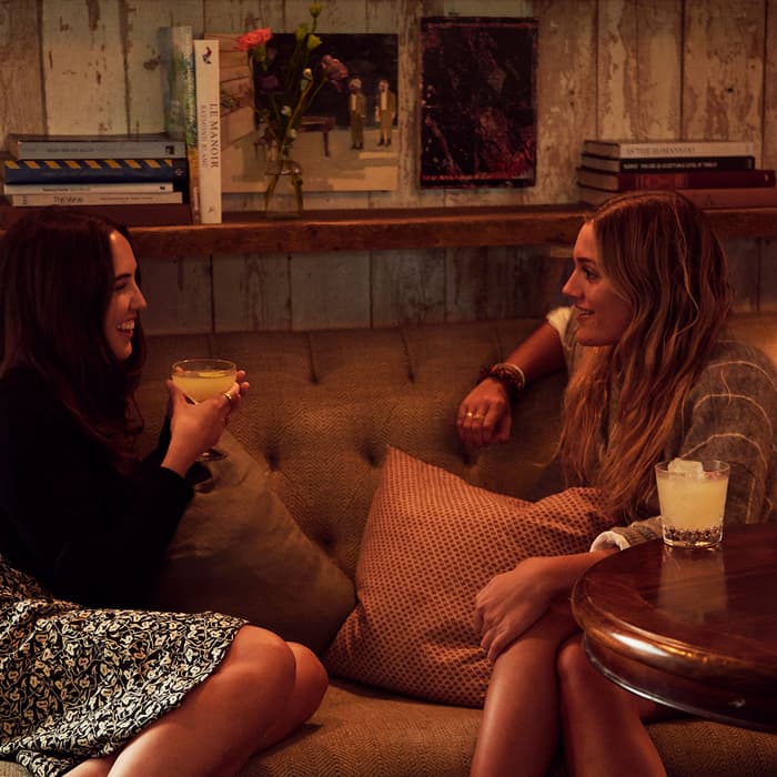 Two women sit on a sofa, chatting and holding drinks. The cozy room features shelves of books, artwork, pillows, a lamp, and a small wooden table with a drink.