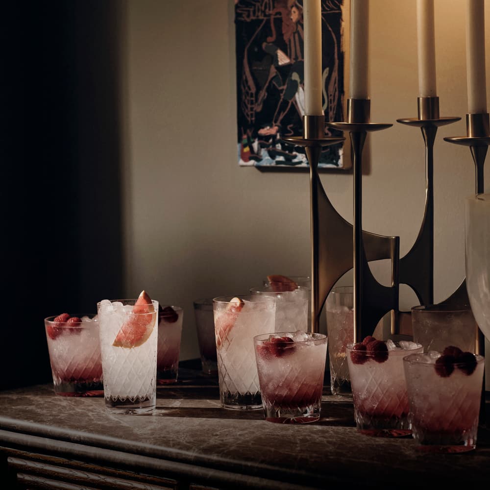 Crystal glasses filled with ice and garnished with grapefruit slices and raspberries sit atop a marble-topped sideboard. Nearby, multiple candles in a modern holder are grouped together.