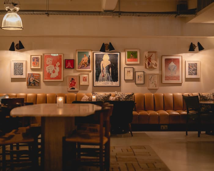 Framed artwork adorns the wall above a tan leather banquette in a dimly lit lounge, featuring wooden tables and chairs, candles, and muted lighting.