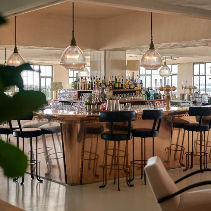 A stylish bar with a polished copper counter, surrounded by black cushioned barstools and under warm pendant lights, sits in a spacious, well-lit room with large windows and modern decor.