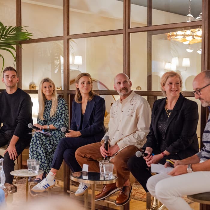 Six individuals are seated in a panel discussion holding microphones, in a warmly-lit room with indoor plants and a glass partition wall. Another person is standing, speaking into a microphone.