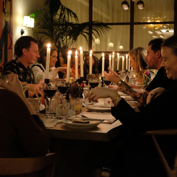 A group of people dining at a long table adorned with lit candles, wine glasses, and ornate cutlery in a warmly lit restaurant with plants and artwork on the walls.