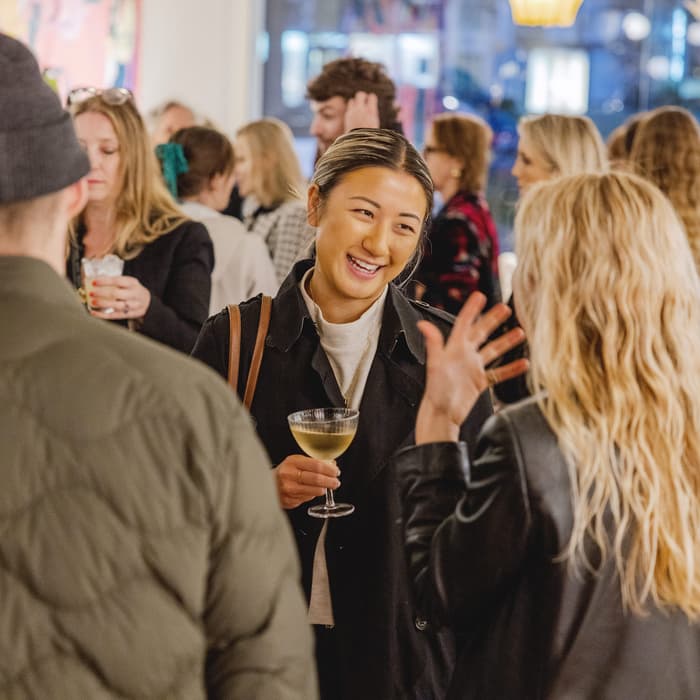 People engaged in animated conversations while holding drinks at a social gathering inside a well-lit room, filled with a crowd and colorful artwork on the walls.