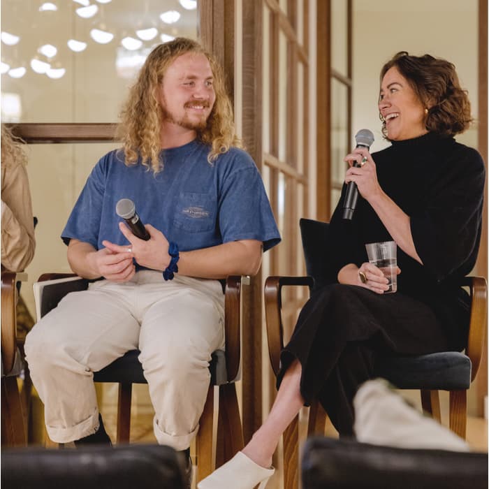 Two people, each holding a microphone, engage in conversation while seated on wooden chairs, inside a well-lit room with glass-paneled walls and modern lighting fixtures.