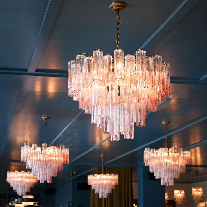 Multiple glass chandeliers hang from a ceiling, illuminating a spacious room with a blue ceiling and walls, and additional chandeliers visible in the background.