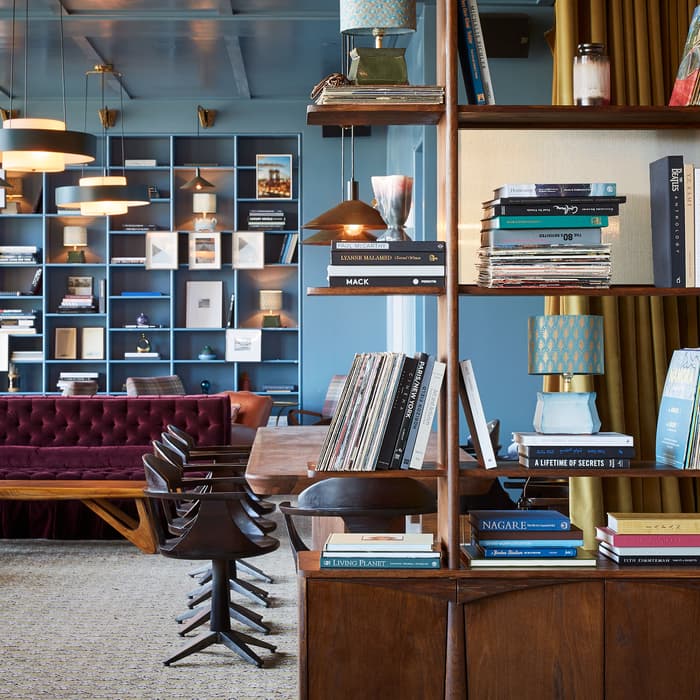 Bookshelves filled with books, lamps, and records. Behind, there are modern hanging lights, a maroon couch, and a large wall of shelves with various decor in a cozy, well-lit lounge.