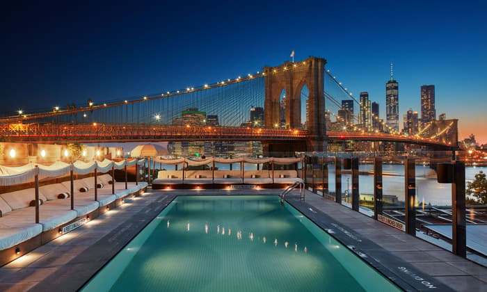 A rooftop swimming pool overlooks a lit-up Brooklyn Bridge and the illuminated Manhattan skyline at twilight. Cushioned lounge chairs with fringed umbrellas line one side of the pool. Text: "4 FEET 3 INCHES DEEP," "NO DIVING."