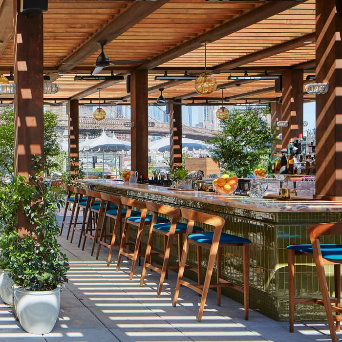 Spacious outdoor bar in sunny, modern setting with wooden beams, numerous potted plants, wooden tables, chairs, and bar stools. A bridge is visible in the background.