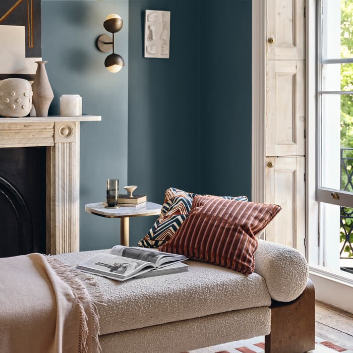 A chaise lounge with pillows and a blanket is placed by a window, next to a small table with a book and glass. A fireplace mantle adorned with vases and artwork is behind.