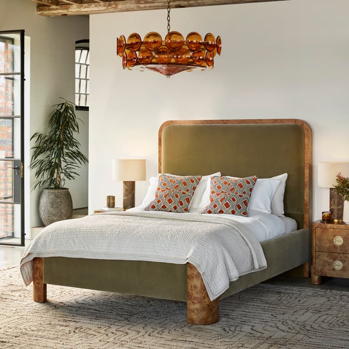 A green bed with white bedding and patterned pillows sits on a patterned rug in a room with exposed wooden beams, a large window, and a brown chandelier.