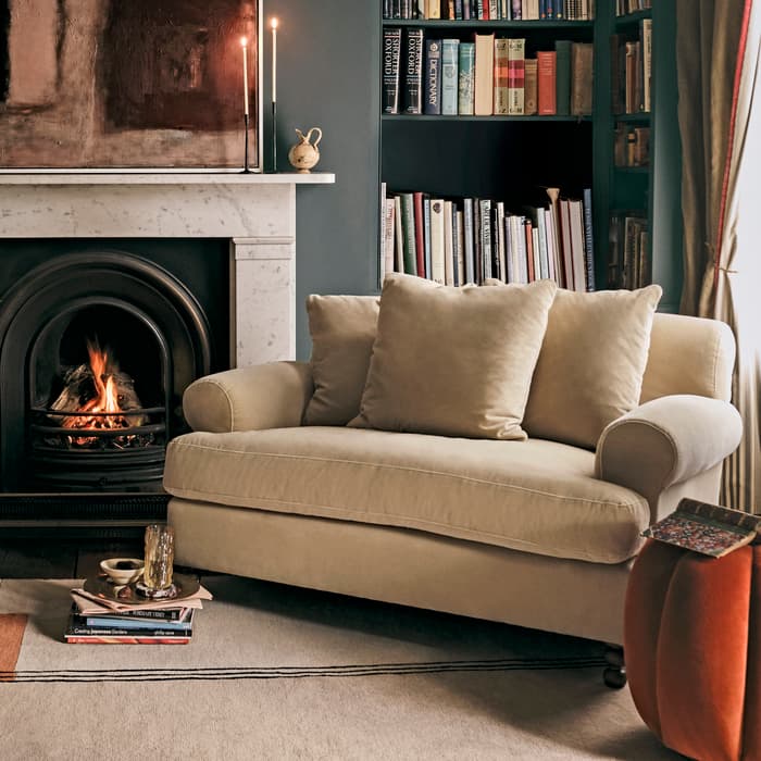 Beige sofa with plush cushions sits before a lit fireplace in a cozy living room, adjacent to a book-filled shelf; candles and a modern painting adorn the mantel.