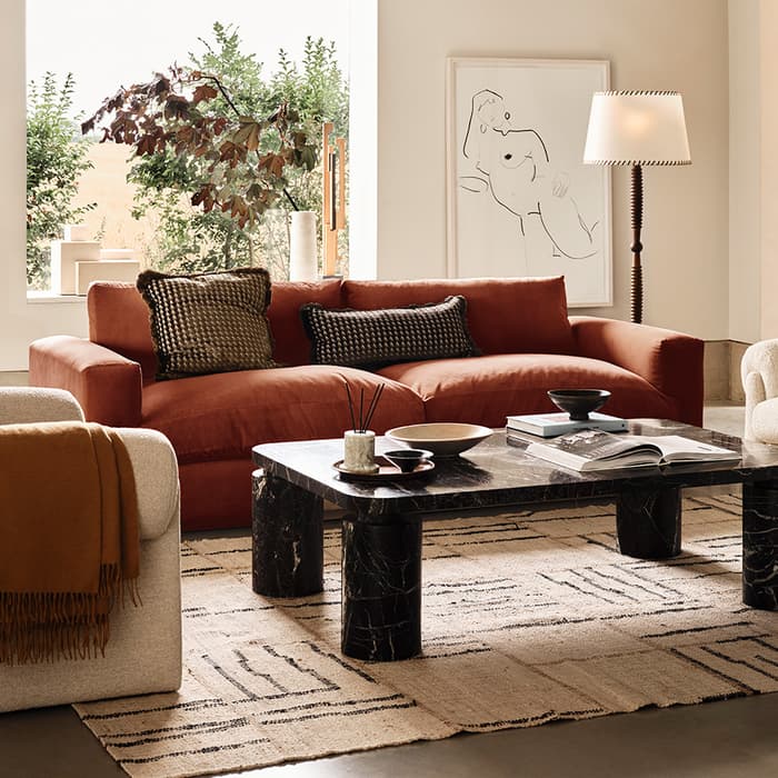 A living room with a rust-colored sofa, a marble coffee table holding books and decor, a beige armchair with a brown throw, a floor lamp, and a large painting, set against a backdrop of greenery through a large window.