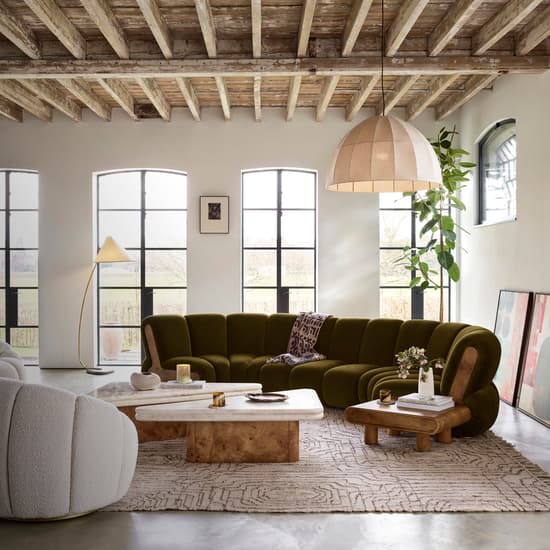 A green sectional sofa and two white plush chairs are arranged around wooden coffee tables in a spacious living room featuring large windows, a rustic wooden ceiling, and modern decor items.