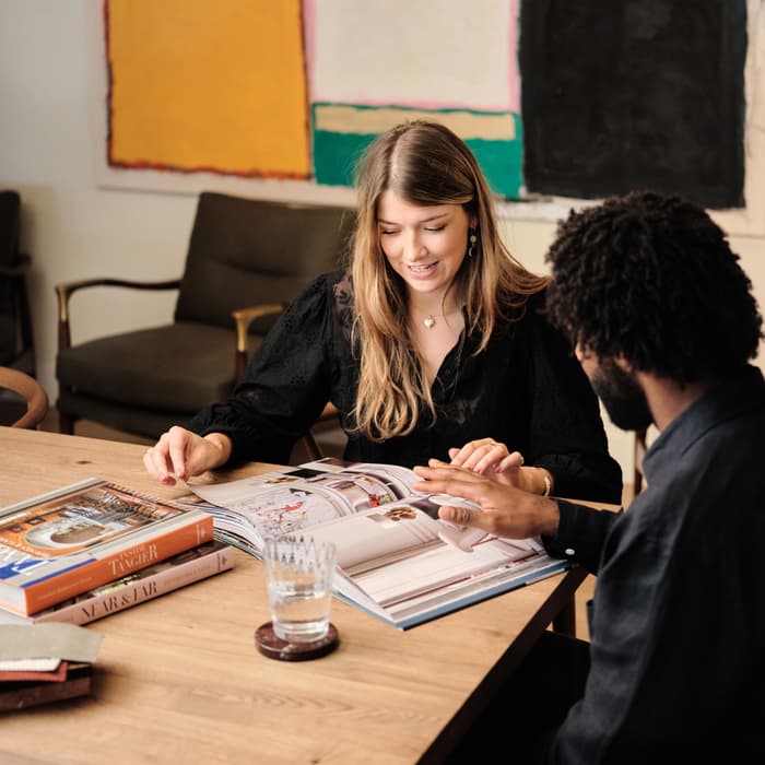 Two people sit at a wooden table, flipping through an open book, with additional books and a glass of water nearby. A colorful abstract painting hangs on the wall behind them.