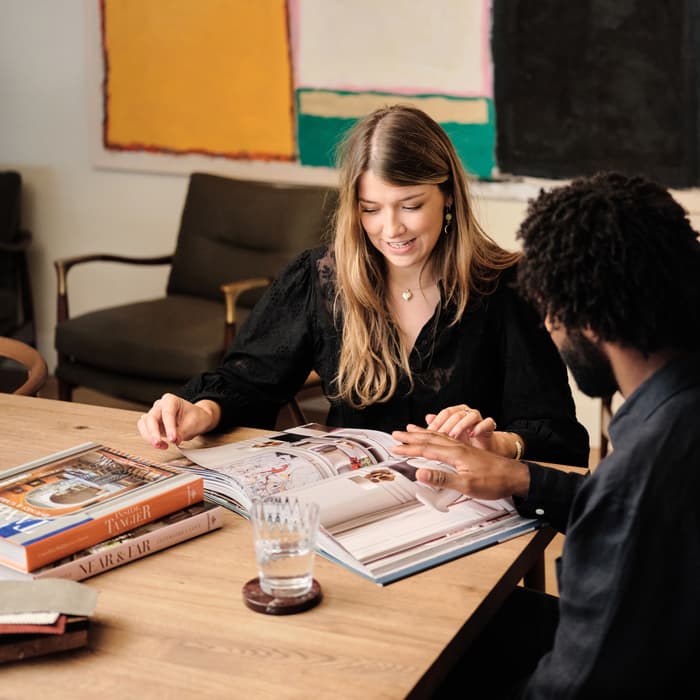 Two people read a book together at a wooden table, surrounded by books and a glass of water, with colorful abstract art on the wall behind them.