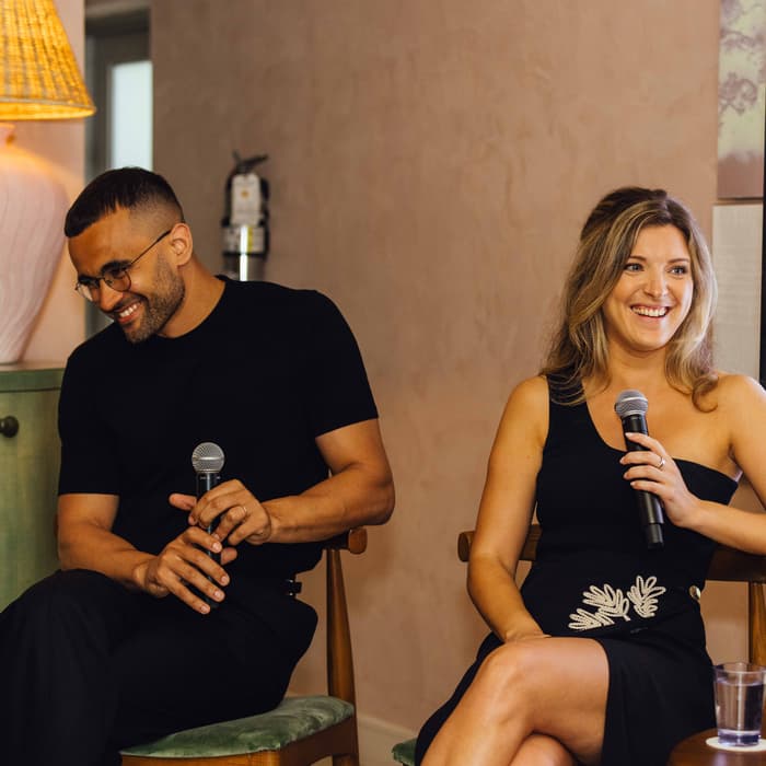 Two people are seated, each holding a microphone, smiling, and engaging in a conversation. The setting includes a side table with a lamp, glass, and books, soft lighting, and a wall-mounted display featuring design ideas labeled "SOHO HOUSE DESIGN TOUR."