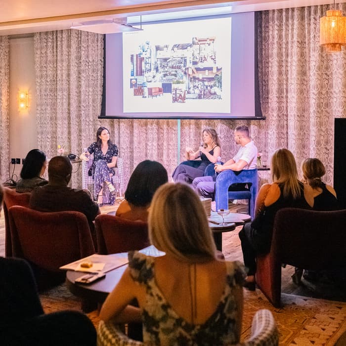 Speakers sit on stage discussing a topic, while an audience listens attentively in a warmly lit room with curtains and hanging light fixtures, a projector screen displays images.